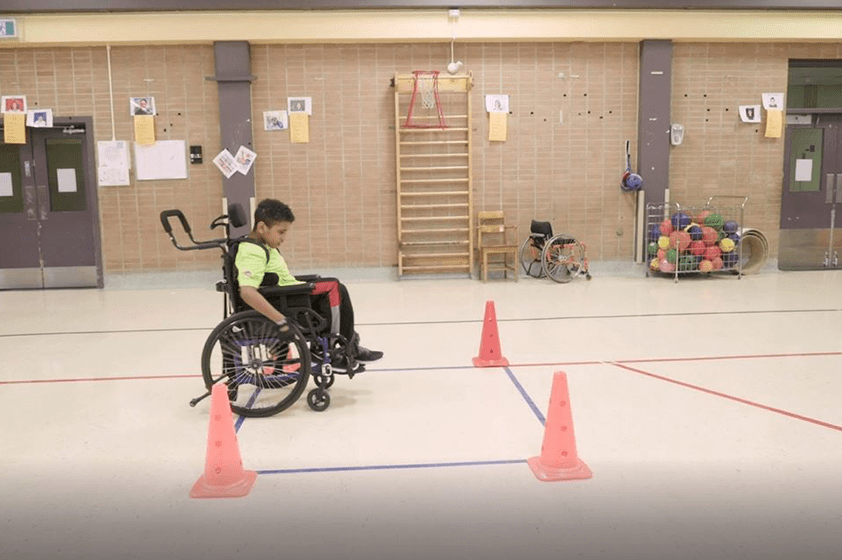 Slalom Une jeune athlète en fauteuil roulant roule entre 4 cônes orange.