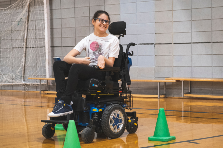 Une fille souriante en fauteuil roulant motorisé se trouve au centre de trois cônes vert.