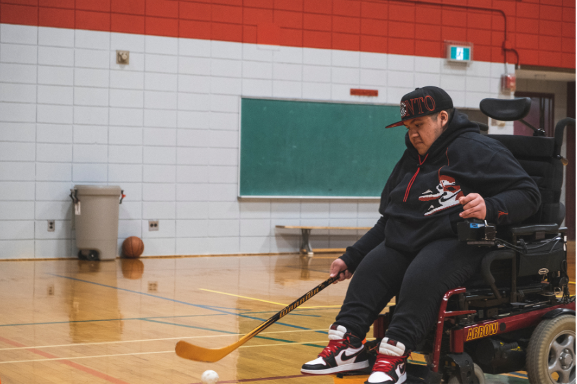 Un jeune en fauteuil roulant motorisé tient un bâton d'hockey de la main droite et dribble avec une balle blanche.