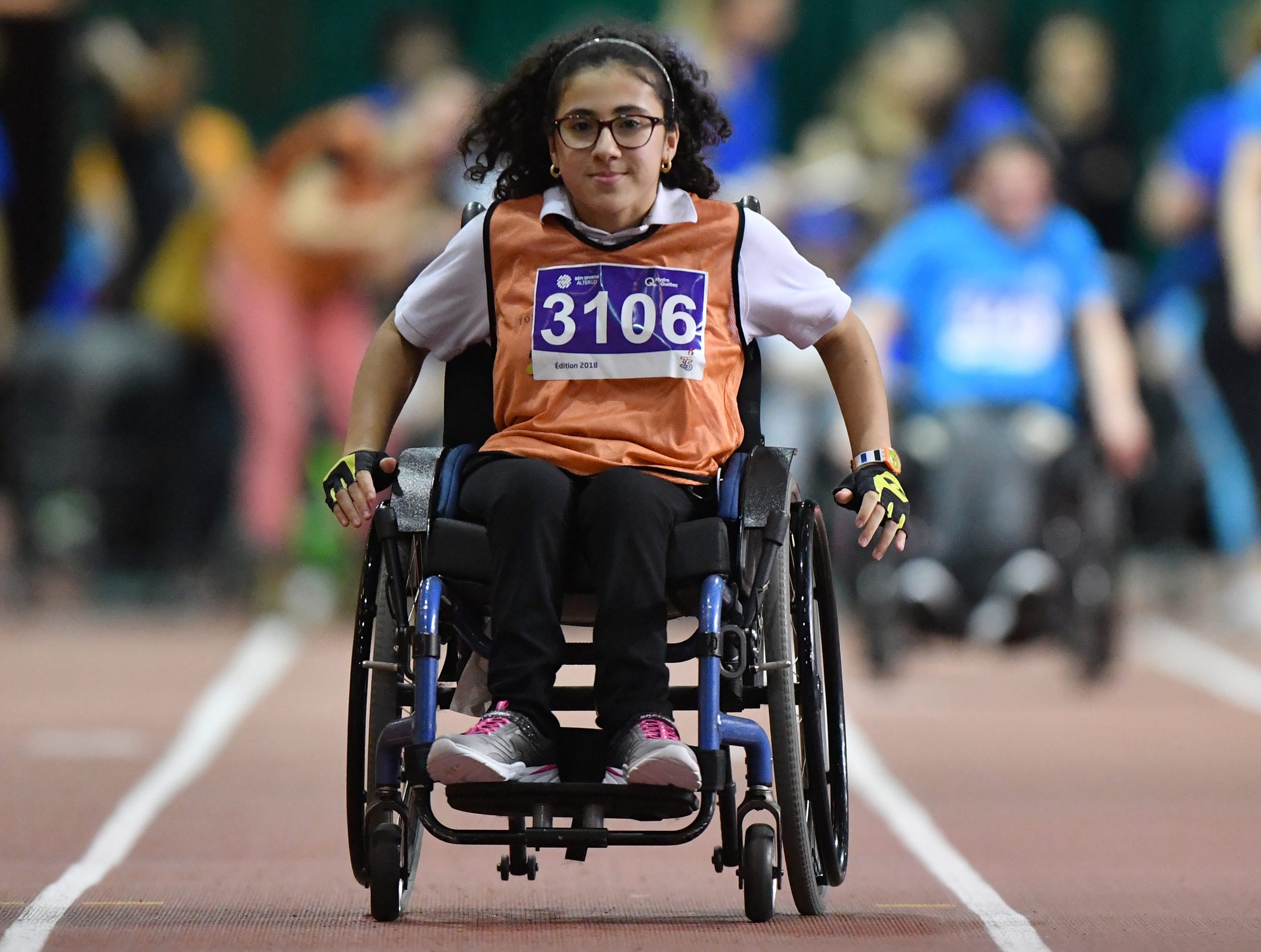 Description de l'image : Au centre, une jeune fille assise sur un fauteuil roulant regarde vers la caméra et compétitionne sur un terrain d'athlétisme lors d'une course.