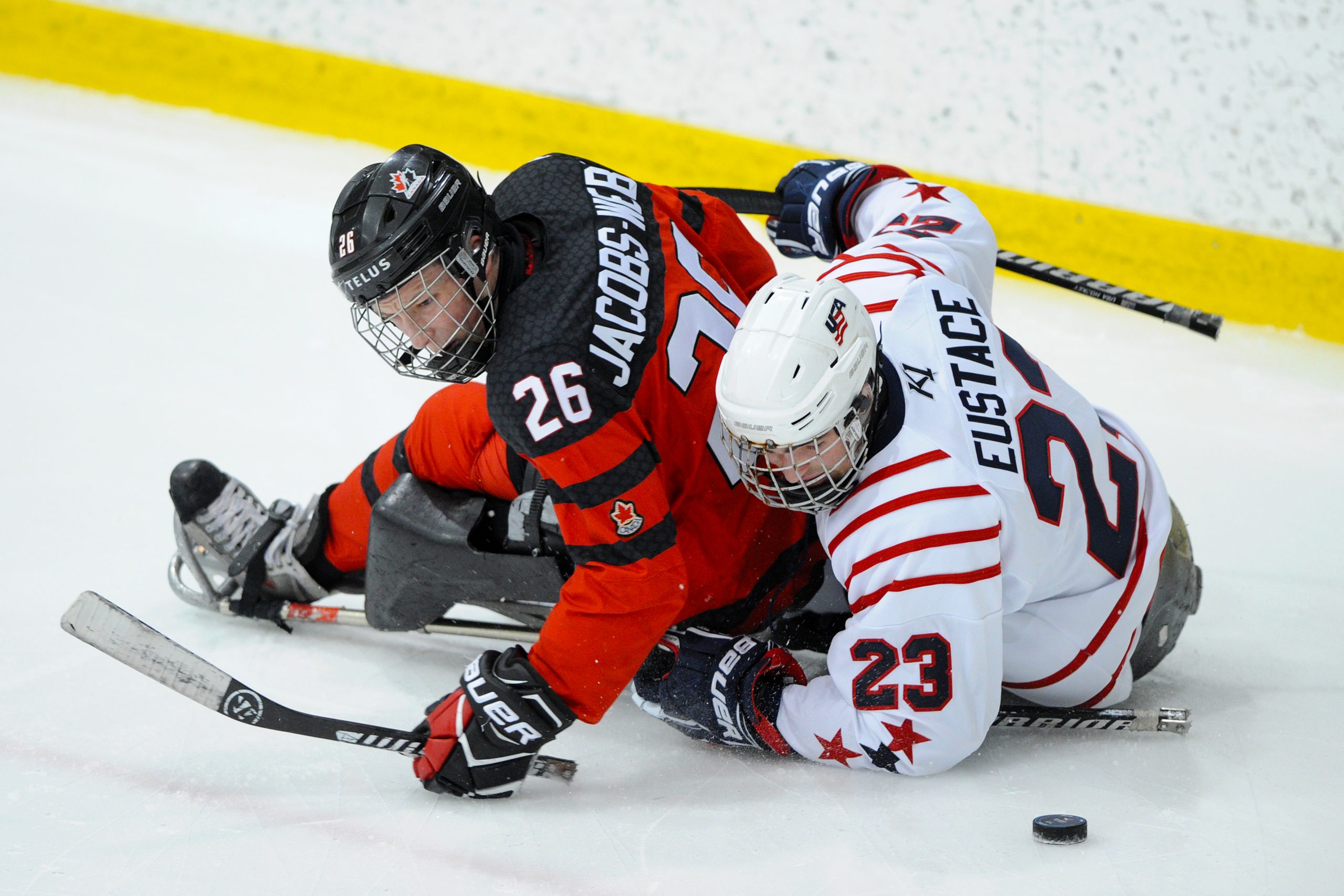 Description de l'image : Un joueur de parahockey de l'équipe canadienne se fait plaquer par un joueur de parahockey de l'équipe américaine. Ils cherchent tous les deux la rondelle, qui est derrière eux.