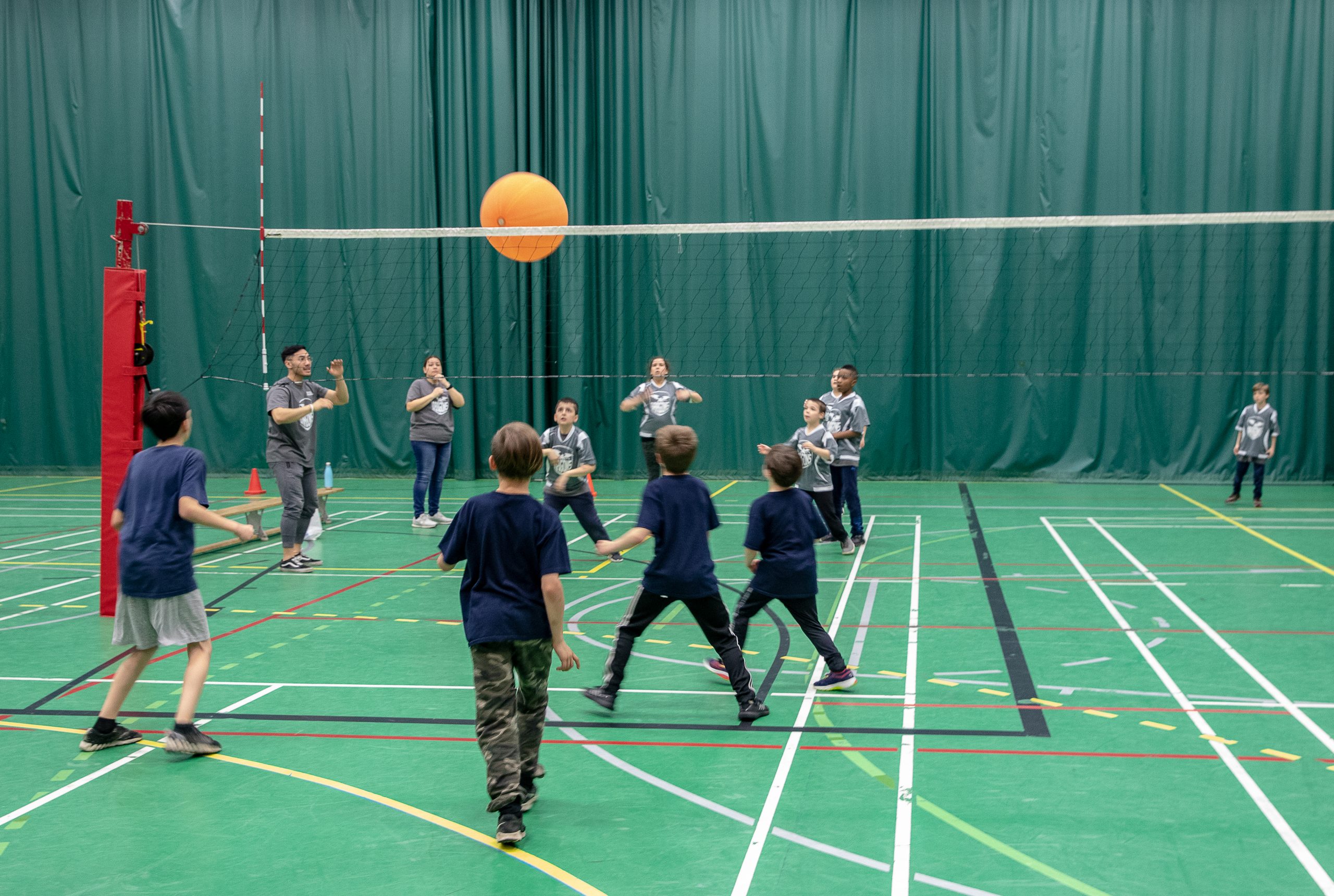 Description de l'image : De jeunes enfants jouent au volleyball dans un gymnase. Le ballon orange traverse le filet.