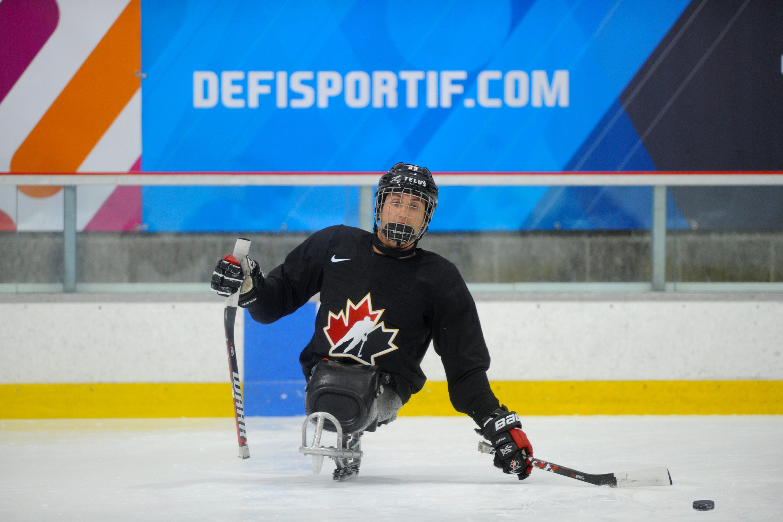 Description de l'image : Au centre, un athlète canadien de parahockey regarde vers la caméra. Son bâton gauche est placé près de la rondelle, qui est sur la glace devant lui.