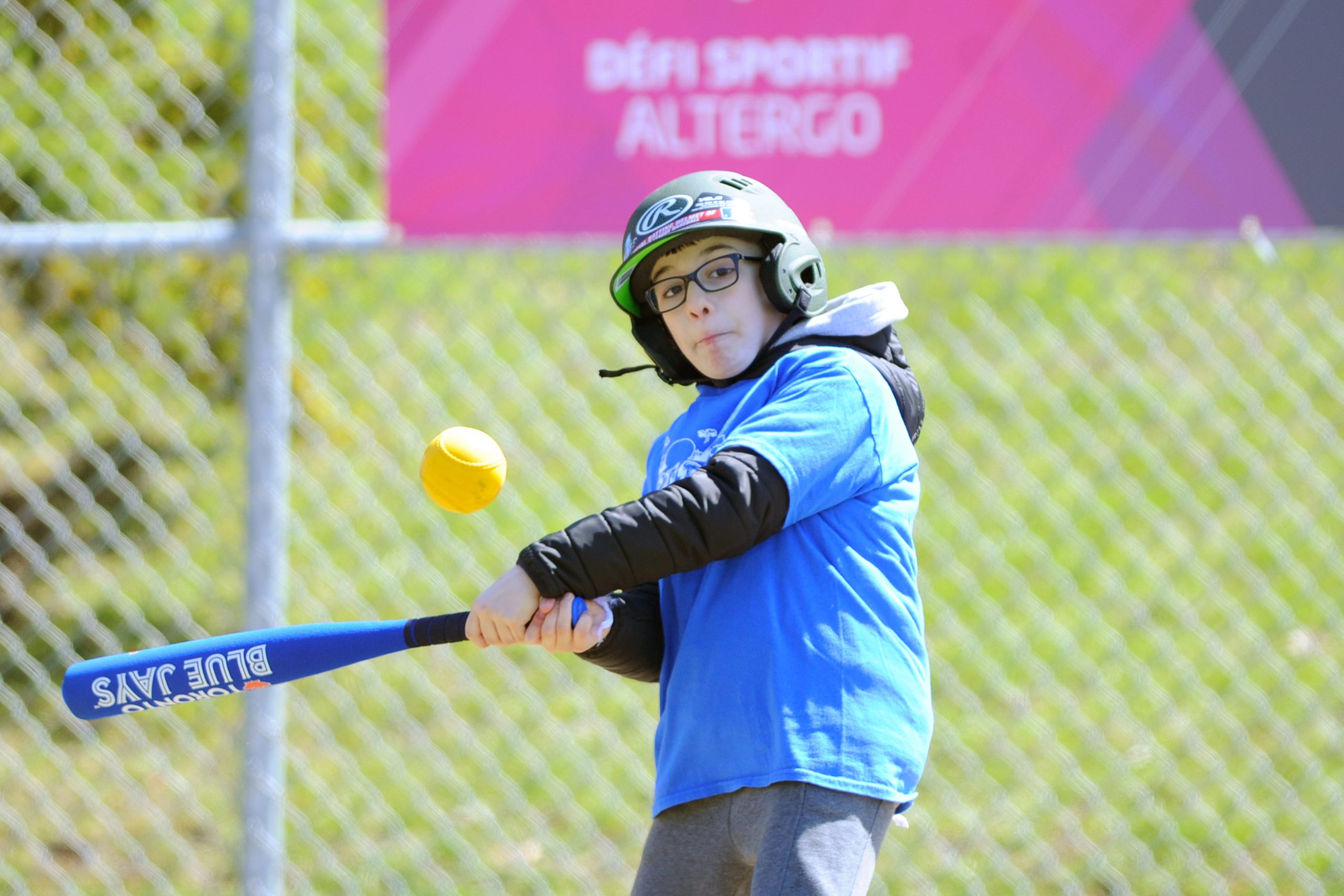 Description de l'image : Au centre, un jeune joueur de baseball s'apprête à frapper la balle jaune qui est dans les airs devant lui.