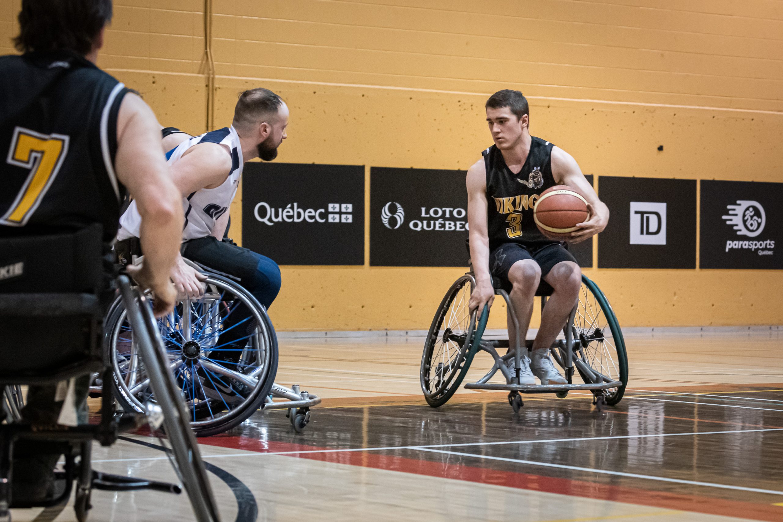 Un joueur de basketball en fauteuil roulant se déplace avec le ballon dans sa main gauche. Il regarde à sa droite, d'où un adversaire vient l'intercepter.