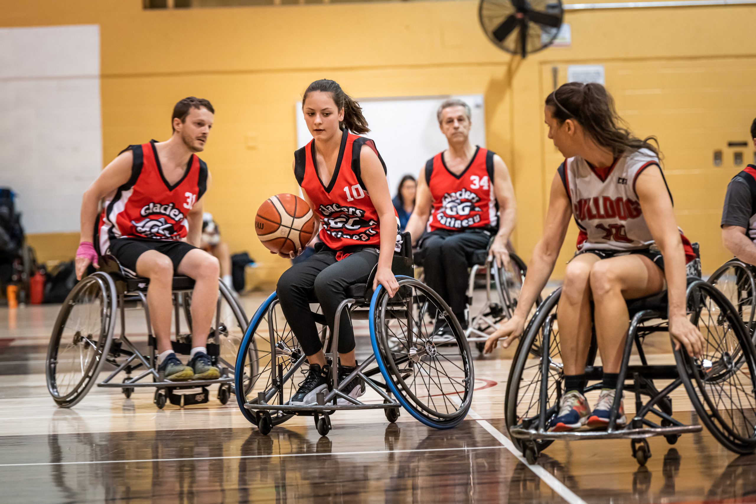 Une joueuse de basketball en fauteuil roulant se déplace avec le ballon dans sa main droite, suivie par deux coéquipiers. Un adversaire est devant elle et la regarde.