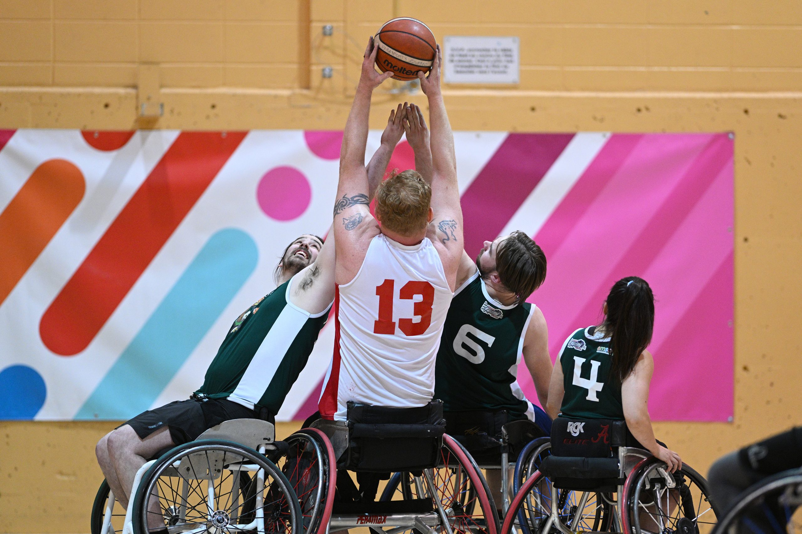 Un joueur de basket-ball en fauteuil roulant tient le ballon du bout de ses bras en haut de sa tête, alors que deux joueurs de l'équipe adverse tentent de lui enlever.